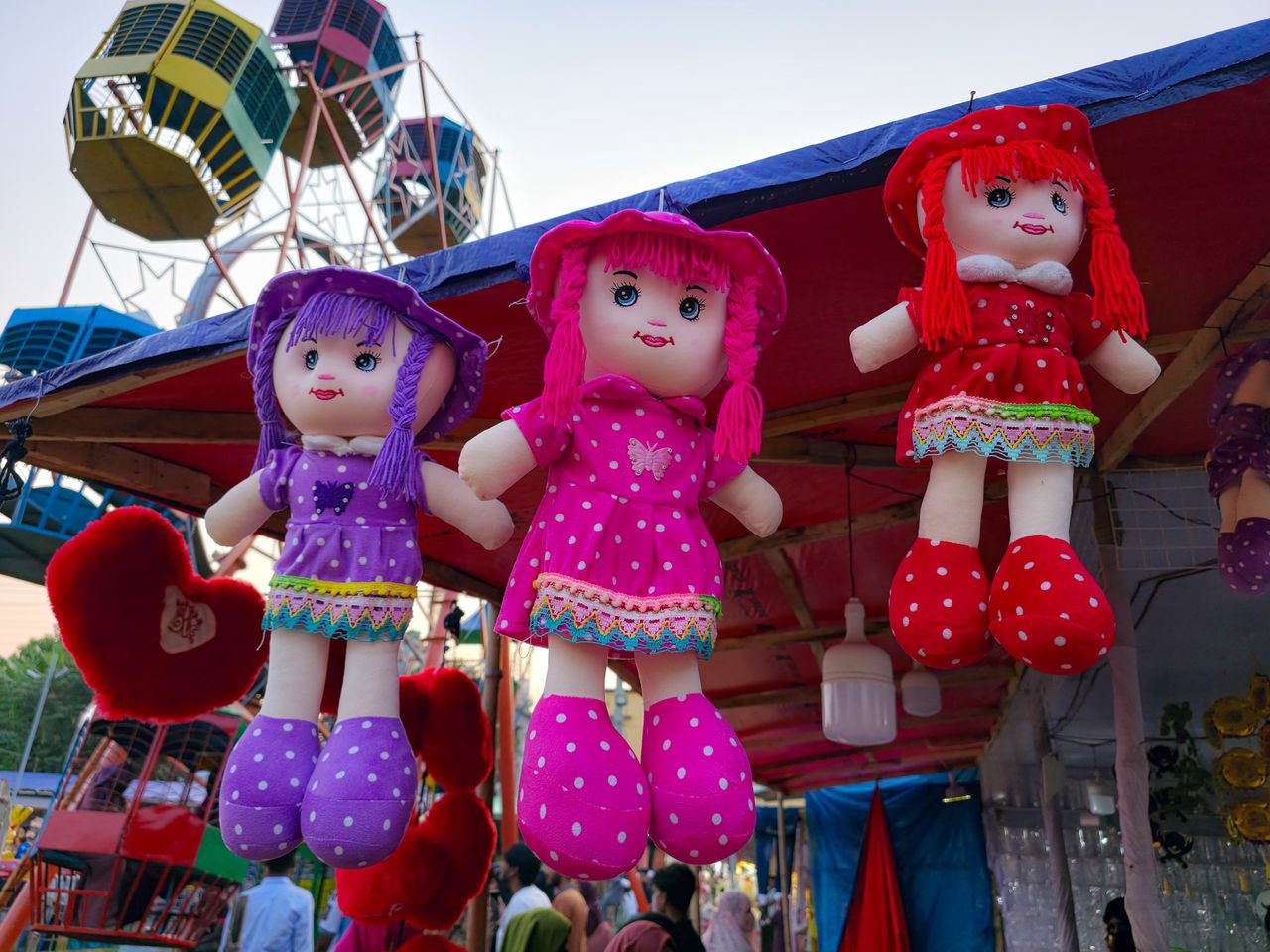 Hanging plush dolls at an outdoor market with a Ferris wheel background.